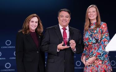 3 smiling people. TCI Representative holding glass award with representatives from the US Dept of State and Concordia